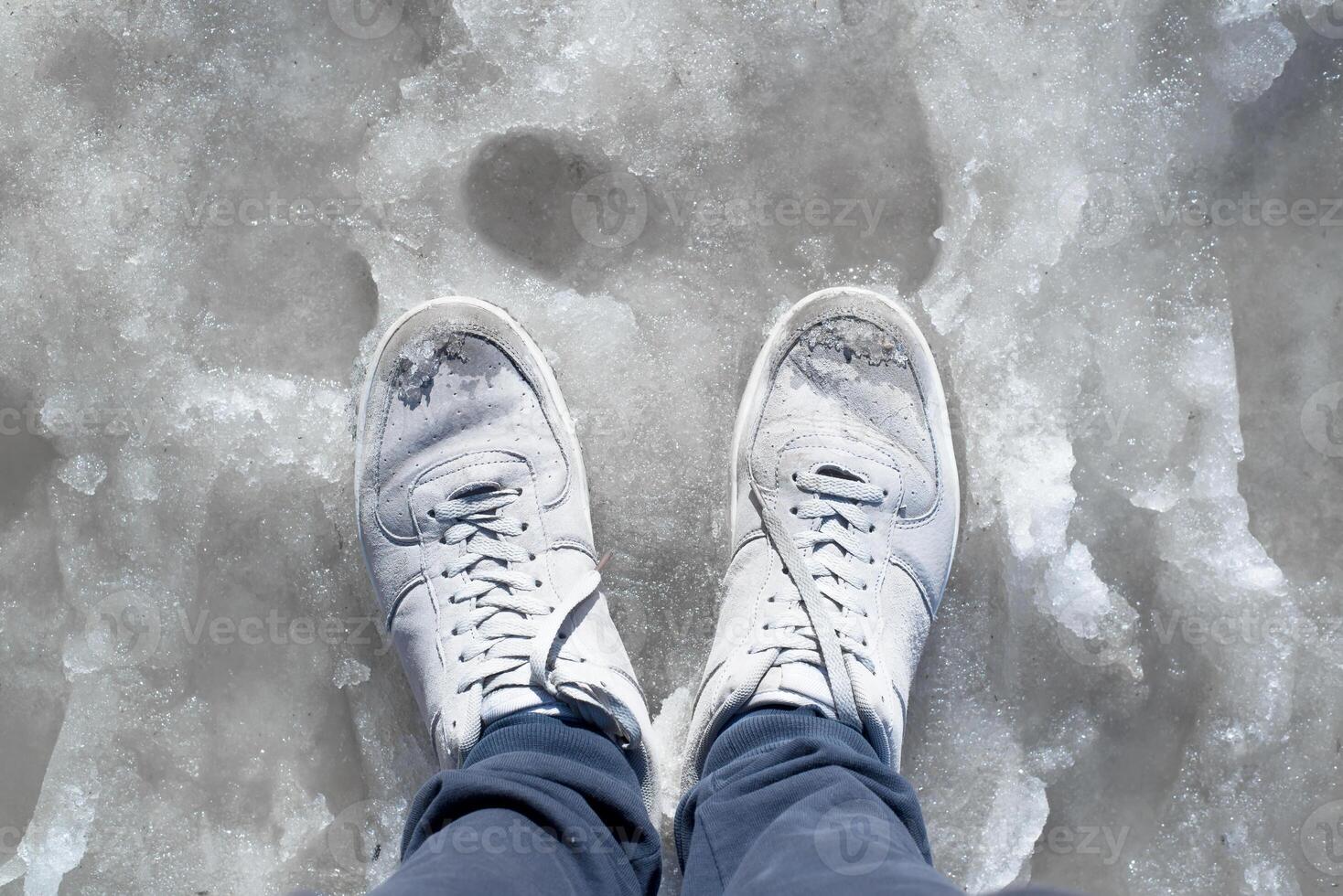 Feet in wet sneakers standing in the melted snow. Puddles, thaw, muddy underfoot on the sidewalk. First-person view photo