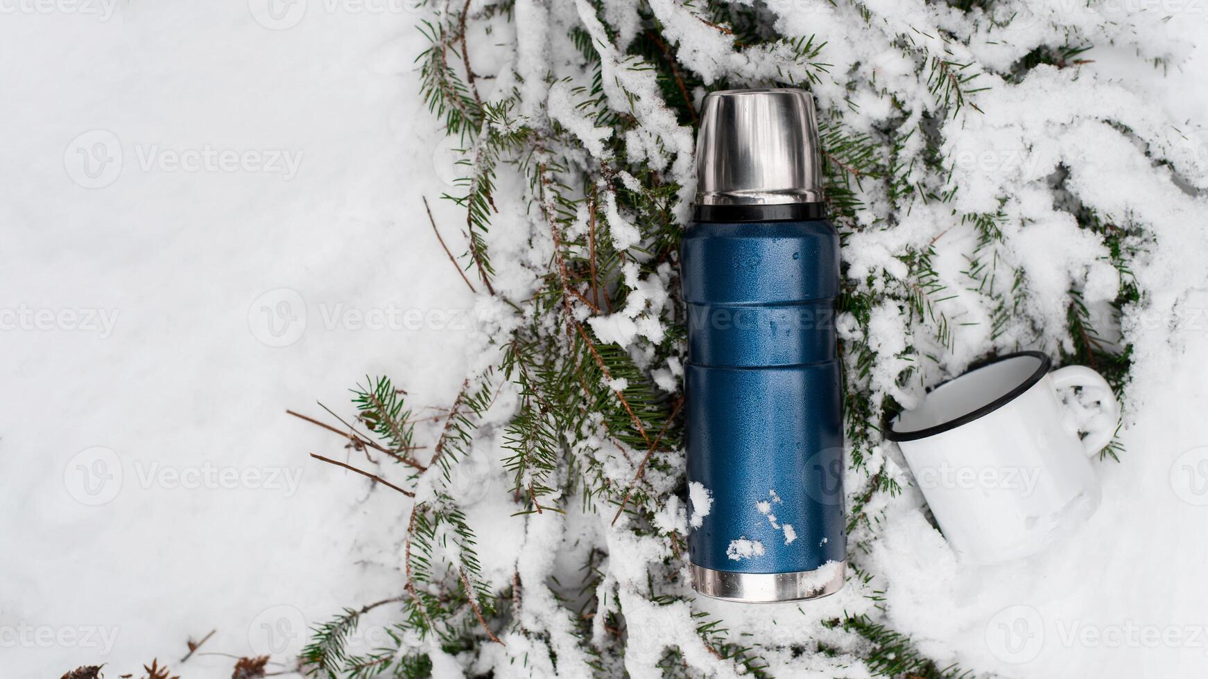 Camping in the winter forest concept. Thermos and iron mug lying on a pine branch in the snow. Top view, copy space. Vacuum flask for travel. photo