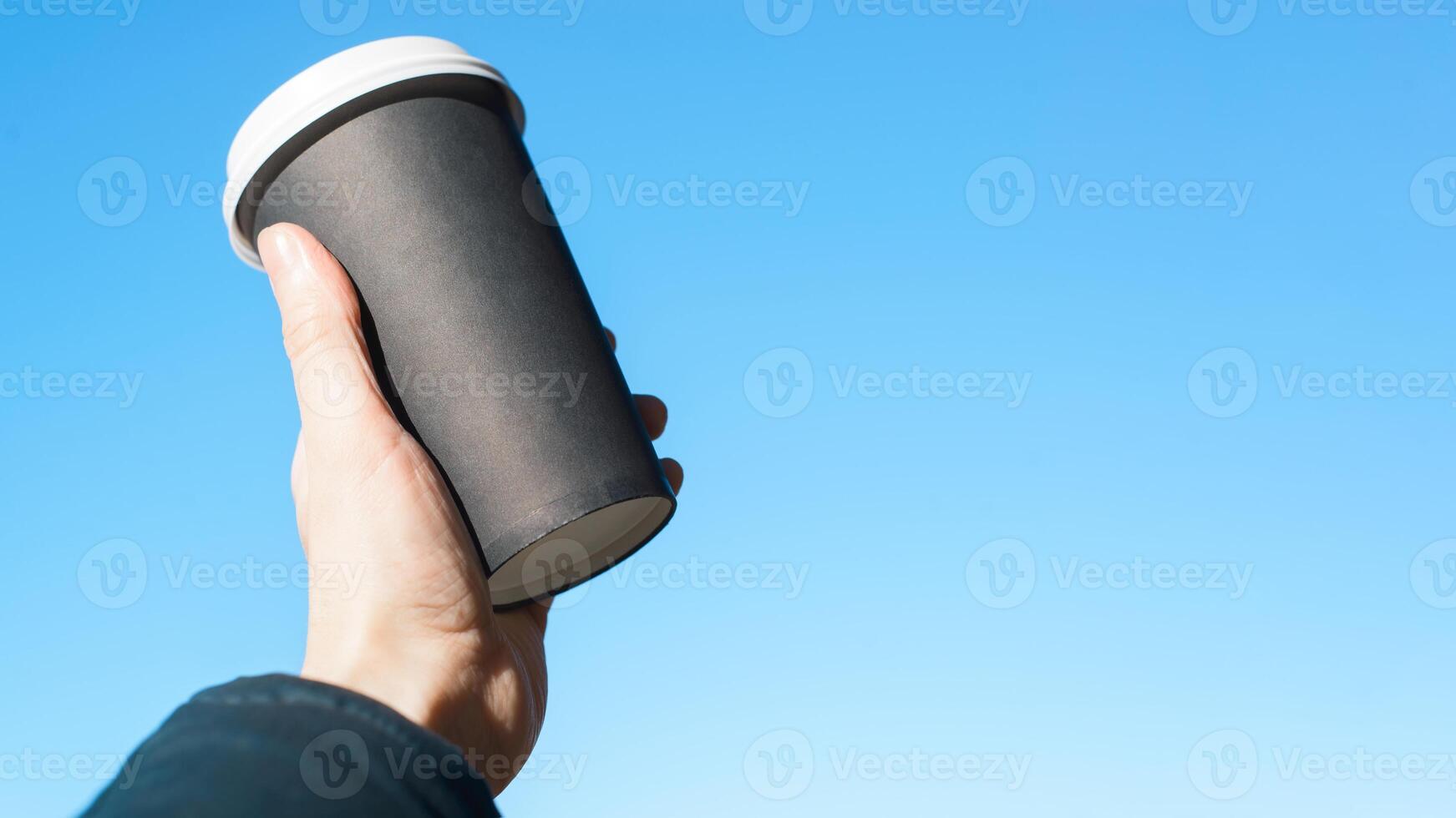A person holding a black coffee cup with a blue sky in the background photo