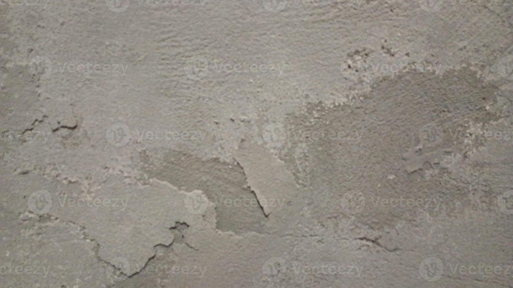 A close-up of a rough, worn-out concrete wall with a gritty texture, featuring cracks and crevices. The color is a dull, muted gray with subtle variations in shade, showing off the uneven surface. photo