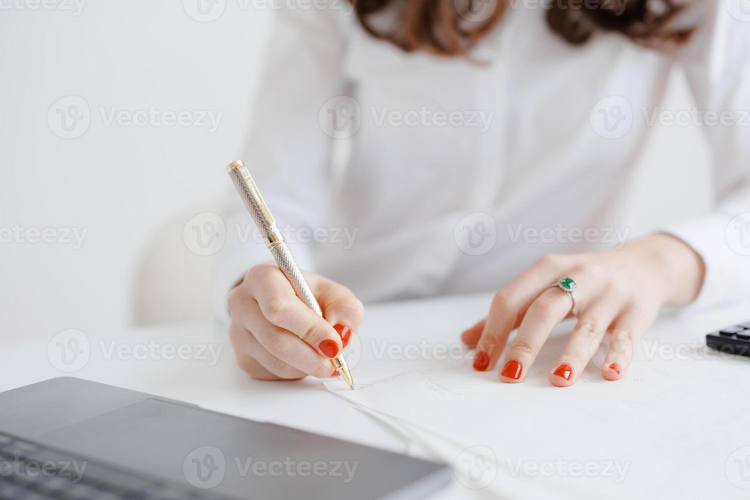 Professional woman writing notes with elegant pen while sitting at desk with laptop and papers photo