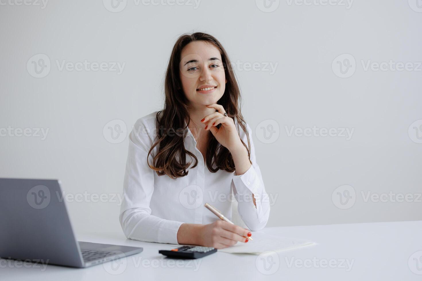 Professional woman smiling while taking notes at a desk with a laptop and calculator in a bright office environment during daytime photo