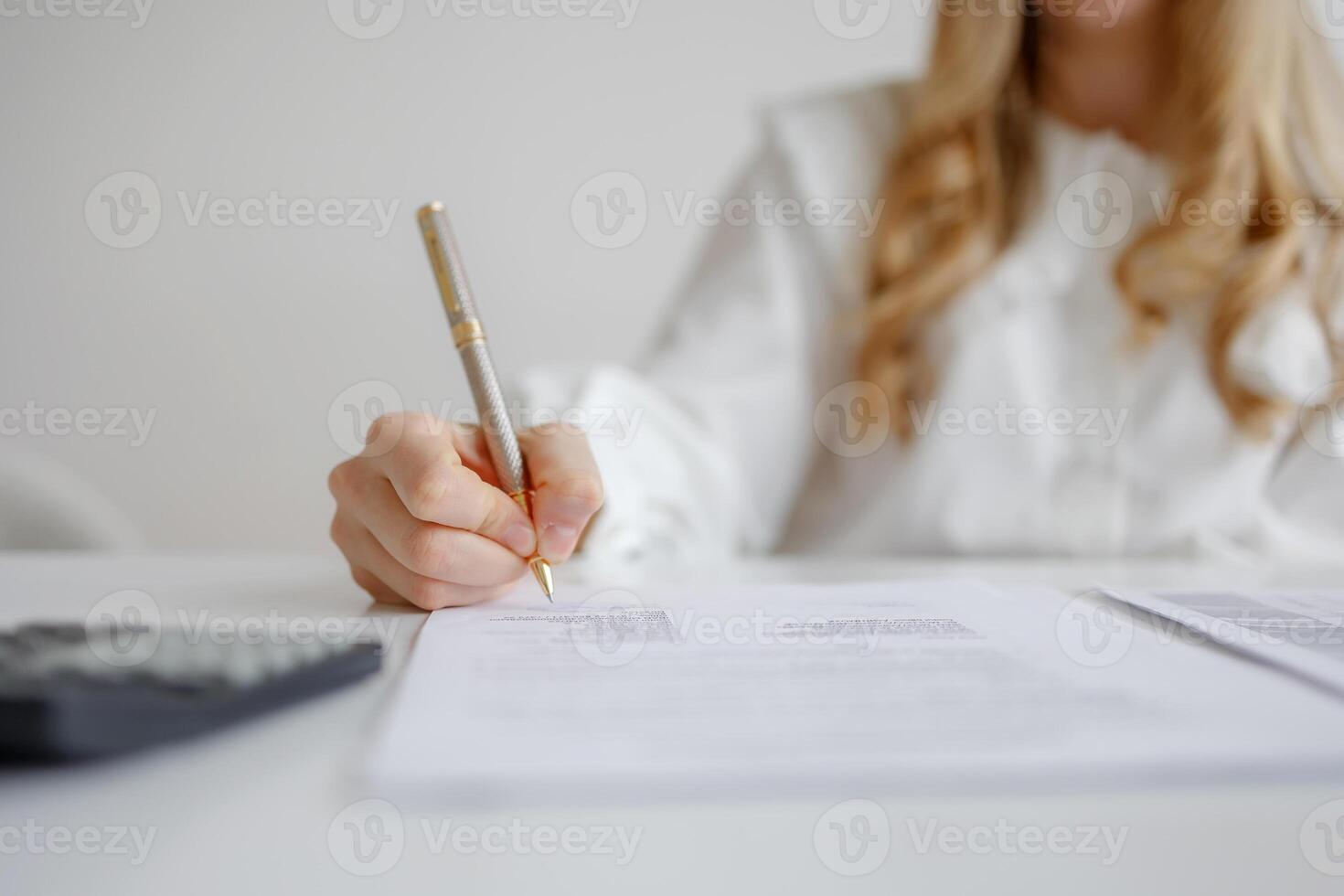 Individual completing paperwork with pen while sitting at desk with calculator and documents in bright workspace photo