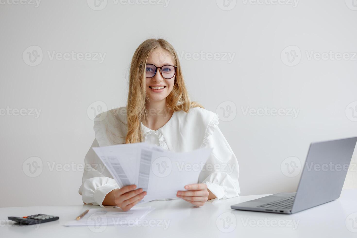 Woman reviewing documents at a desk with a laptop in a bright office environment during the day while smiling photo