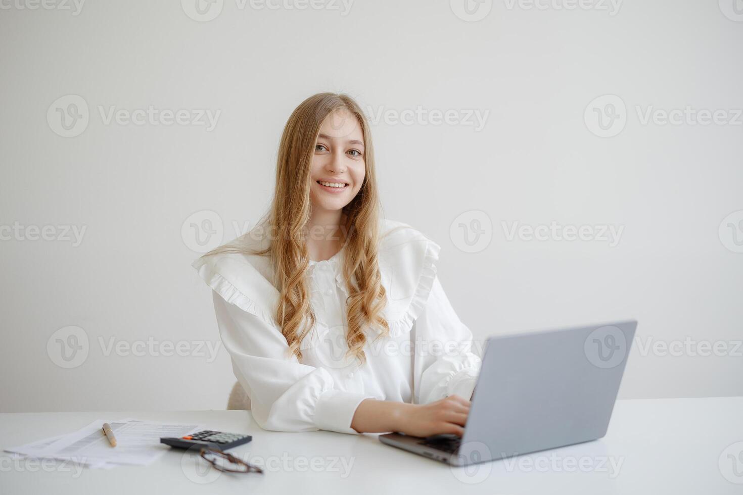 Young woman working on a laptop in a bright, minimal office while smiling towards the camera photo
