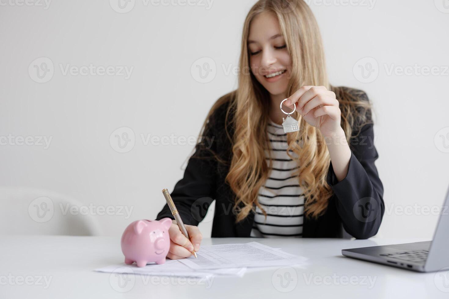 Young woman completing paperwork for new home purchase while holding house key and sitting next to piggy bank photo