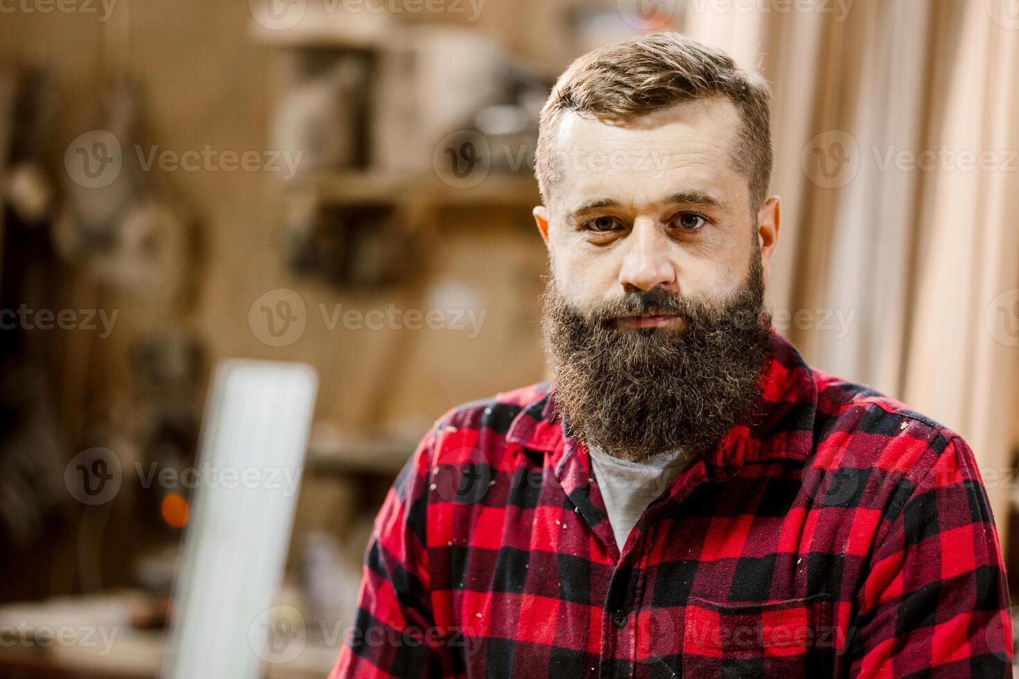Skilled craftsman with beard standing in workshop surrounded by tools and wood materials photo
