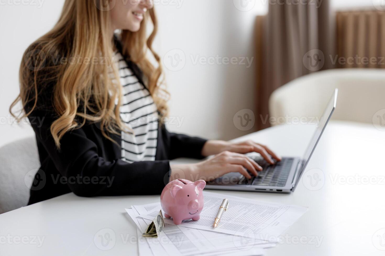 Woman working on laptop at desk with documents and pink piggy bank in modern office setting during daytime photo