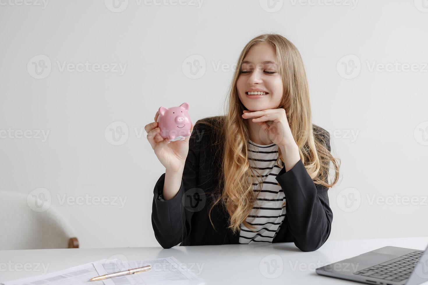 Woman holding piggy bank in modern office while smiling and looking satisfied with financial planning photo