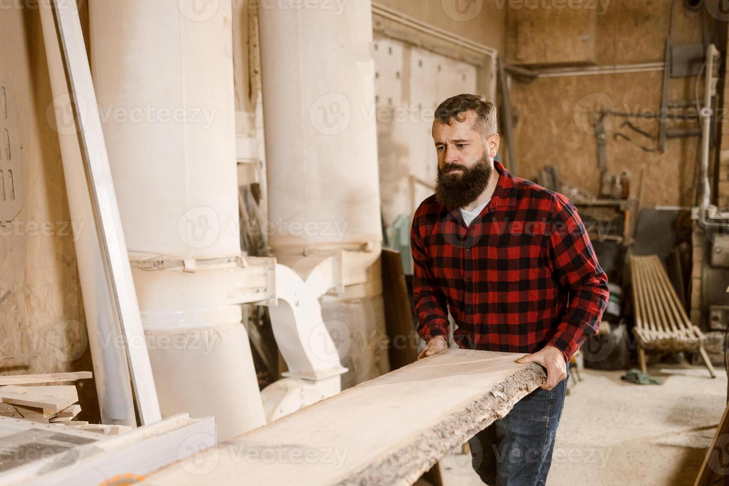 Man transporting a heavy wooden plank inside a workshop during daylight hours photo