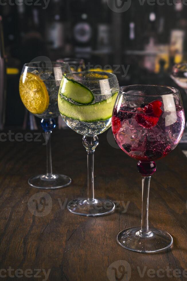 Three elegant gin tonic cocktails with garnishes of orange, cucumber, and red berries on a rustic wooden bar, with a blurry bar background and moody lighting, prfect for mixology or drink concepts. photo