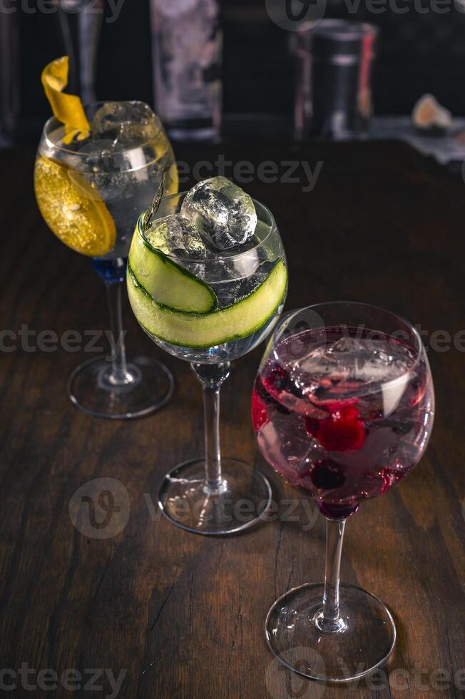 Three elegant gin tonic cocktails with garnishes of orange, cucumber, and red berries on a rustic wooden bar, with a blurry bar background and moody lighting, perfect for mixology drink concepts. photo