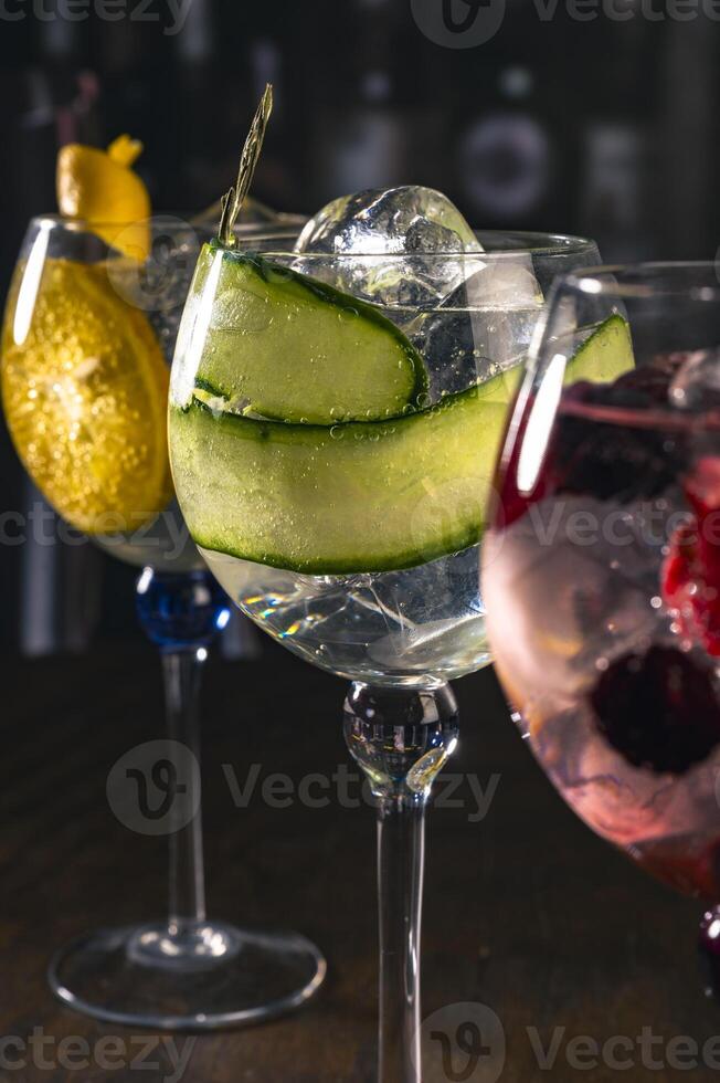 Three stylish cocktails served in large glasses with orange, cucumber and red berries, standing on a wooden surface in a moody bar ambiance, great for concepts of drinks, nightlife or bar aesthetics. photo