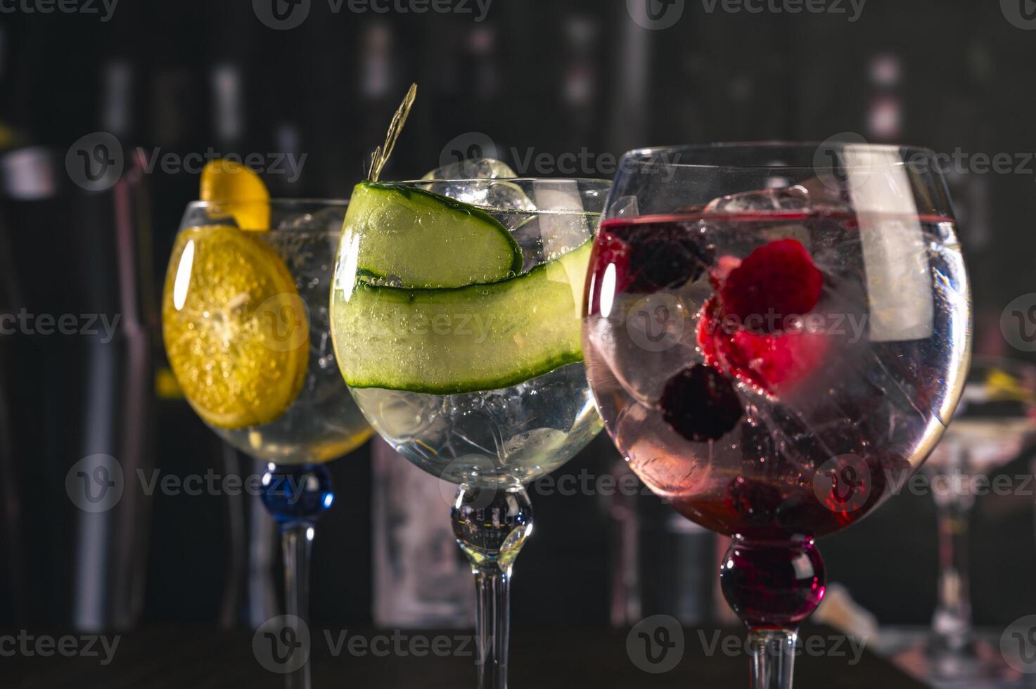 Three stylish cocktails served in large glasses with orange, cucumber and red berries, standing on a wooden surface in a moody bar ambiance, great for concepts of drinks, nightlife or bar aesthetic. photo