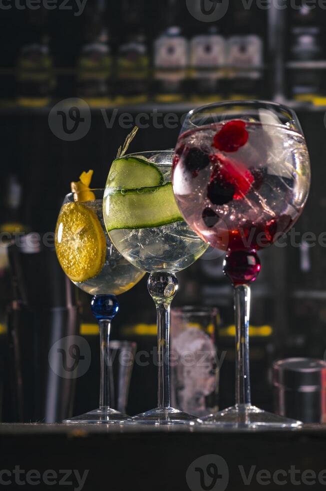 Three elegant gin tonic cocktails with garnishes of orange, cucumber, and red berries on a rustic wooden bar, with a blurry bar background and moody lighting, perfect fr mixology or drink concepts. photo