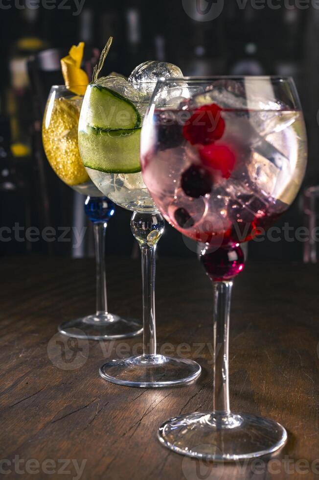 Three elegant gin tonic cocktails with garnishes of orange, cucumber, and red berries on a rustic wooden bar, with a blurry bar background and moody lighting, for mixology or drink concepts. photo
