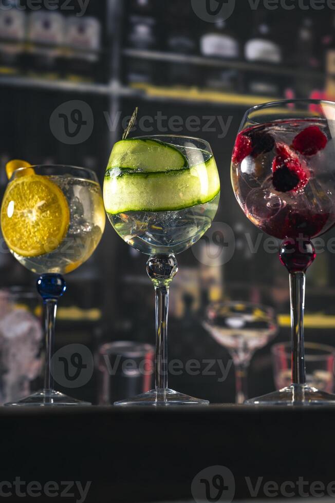 Three elegant gin tonic cocktails with garnishes of orange, cucumber, and red berries on a rustic wooden bar, with a blurry bar background and moody lighting, perfect for mixology or drink concep photo
