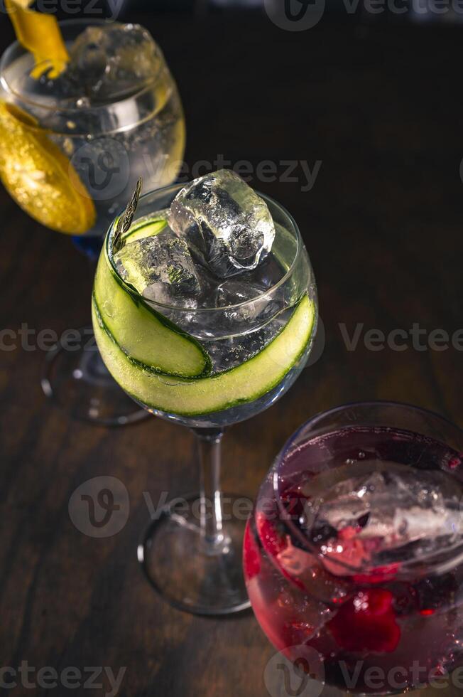 Three elegant gin tonic cocktails with garnishes of orange, cucumber, and red berries on a rustic wooden bar, with a blurry bar background and moody lighting, perfect for mixology r drink concepts. photo