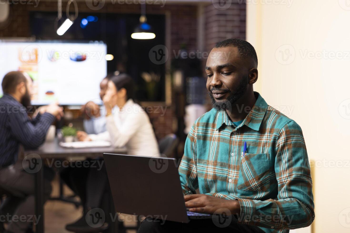 African american entrepreneur reviews financial planning data on device as startup team collaborates in background. Portrait of businessman with laptop, checking emails and planning his schedule. photo