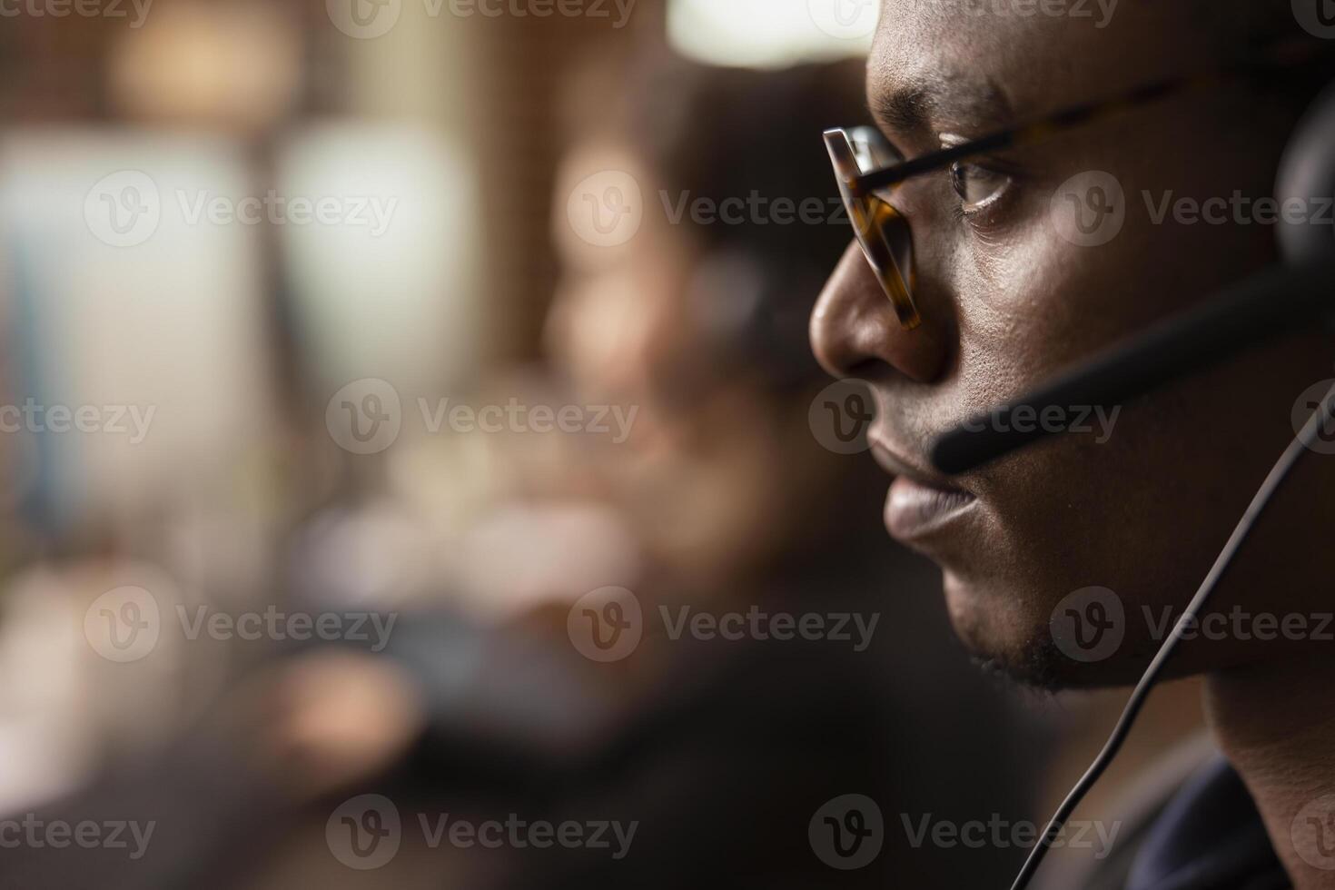 Extreme close up of a black man wearing spectacles, communicating with clients at an online support service office. African American male agent wearing a headset, virtually assisting customers. photo