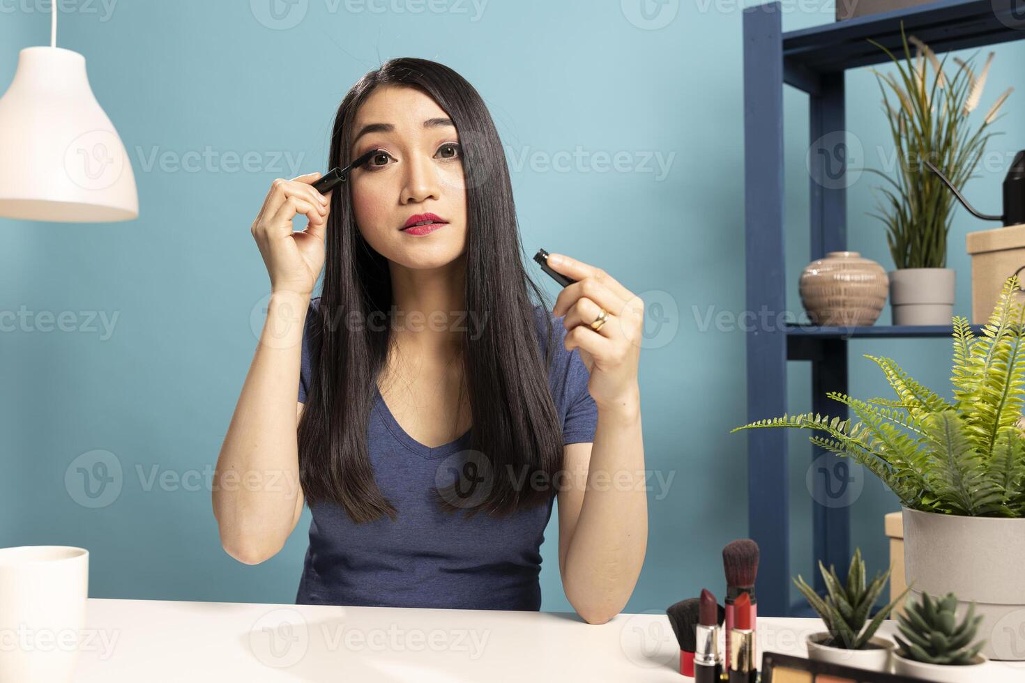 Beautiful woman using cosmetic product while recording a makeup tutorial for online audience. Asian female vlogger seated at desk, demonstrating how to apply mascara during her live broadcast. photo