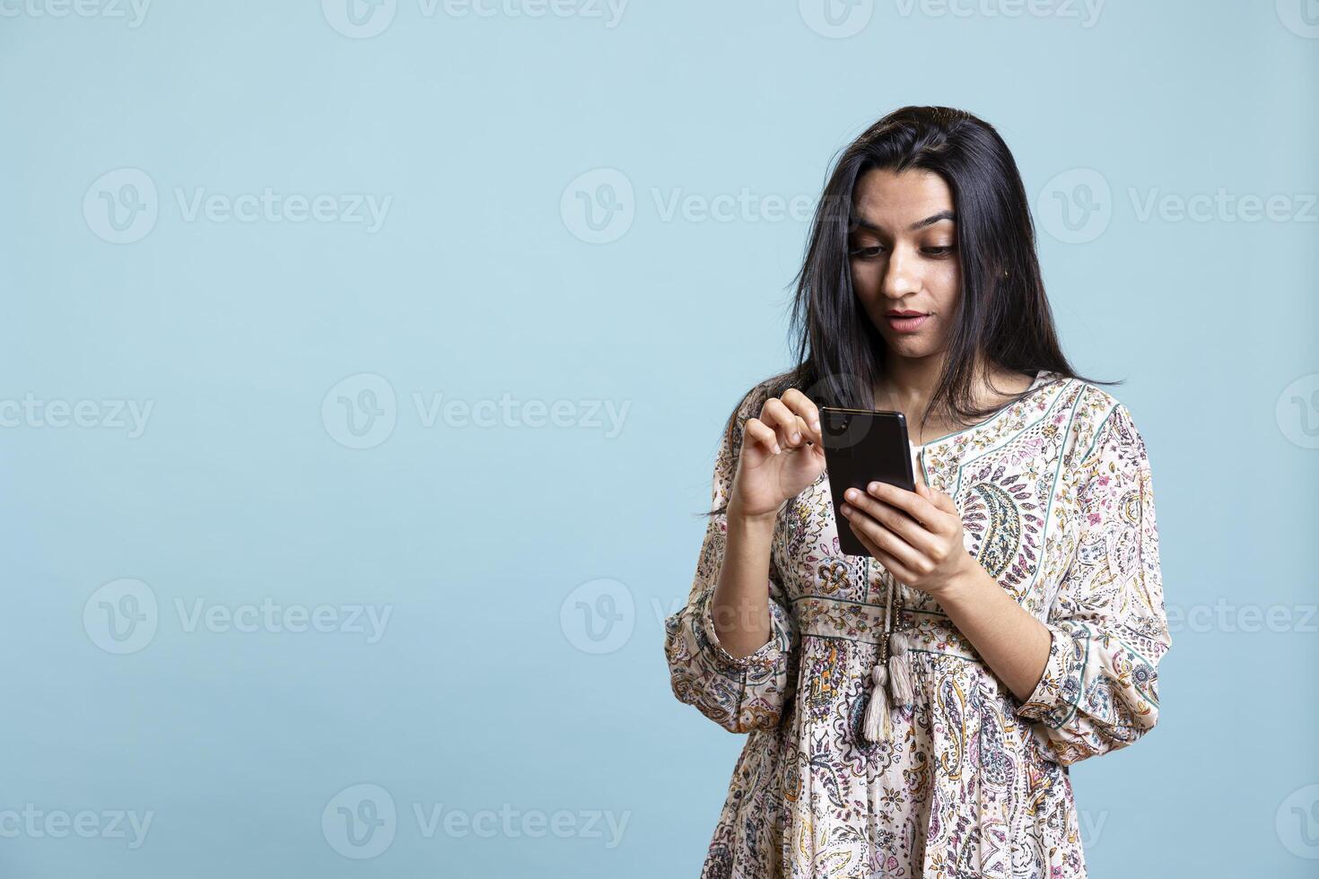 Indian woman happily scrolling on phone to check social media feed, amazed by something. Young relaxed person browsing the internet websites, texting friends against studio background. photo