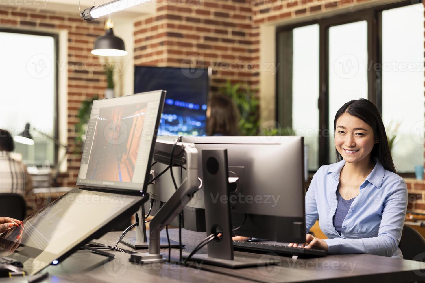 Portrait of asian businesswoman sitting at table and happily working on her computer in modern workspace. Cheerful female professional with her hands on keyboard, typing and looking at camera. photo