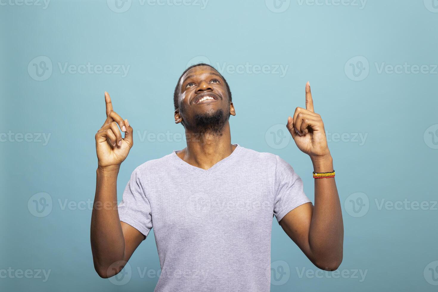 Cheerful african american man pointing upward, looking above as if highlighting a message. Happy male individual in a casual t shirt against a blue background, perfect for an advertisement. photo