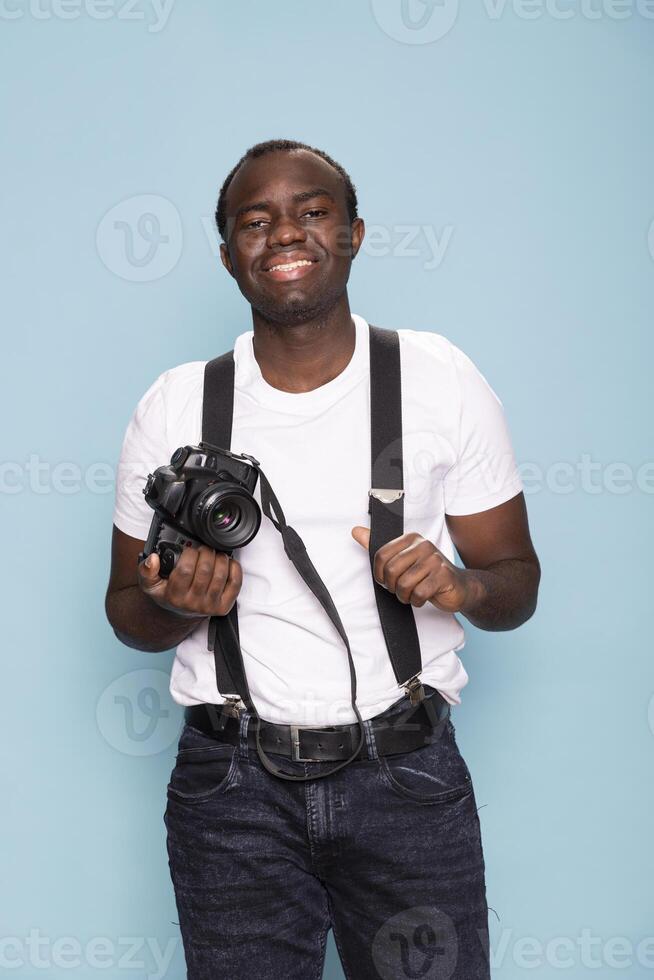 Young man in suspenders and a casual white t shirt holding a professional camera in blue background studio. His warm smile and relaxed posture conveys a welcoming and engaging presence. photo