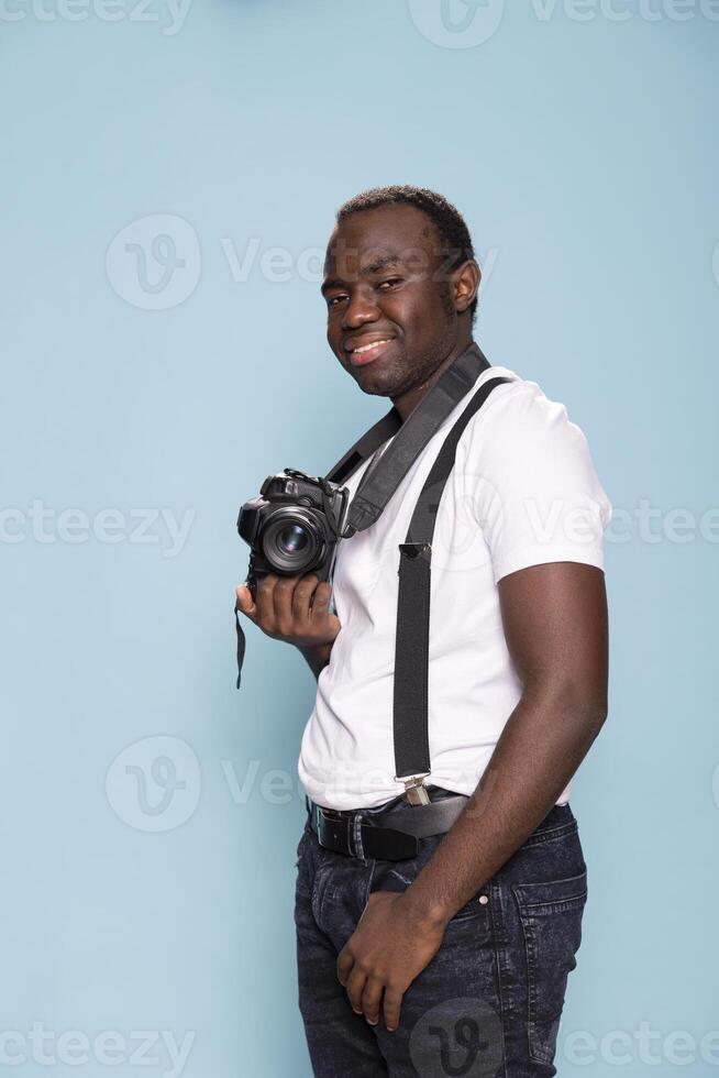 Black man poses with his camera, dressed in casual outfit with suspenders. Portrait of male photographer standing against a blue background. Perfect for advertising, branding, and promotional content. photo