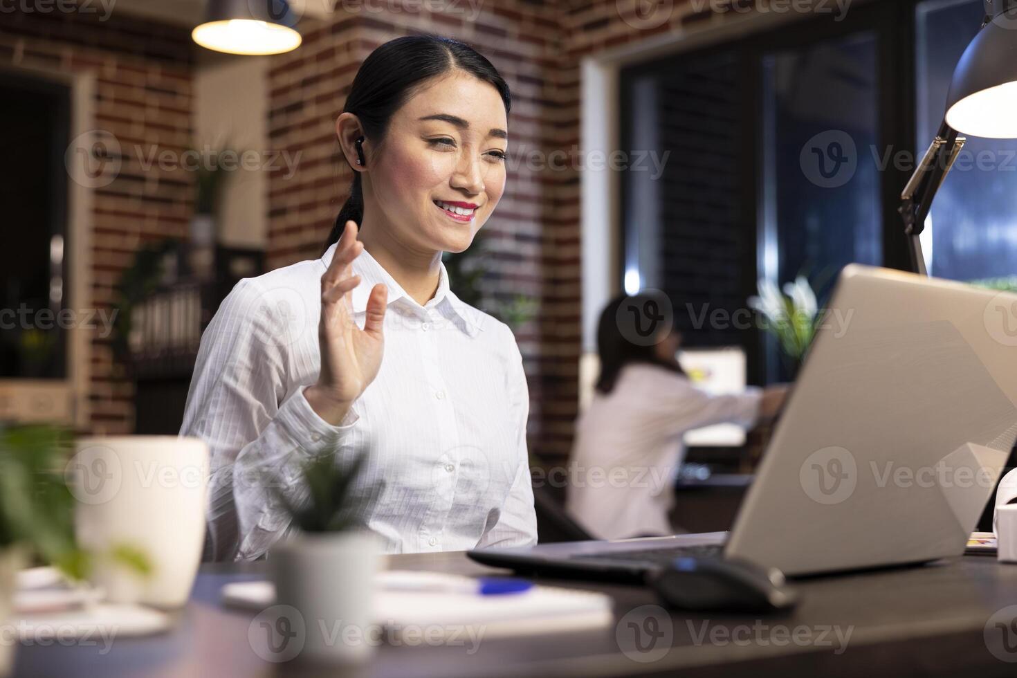 Smiling female manager waving at laptop screen, attending videoconference call with potential client. Asian woman entrepreneur using her personal computer for a virtual meeting with business partners. photo