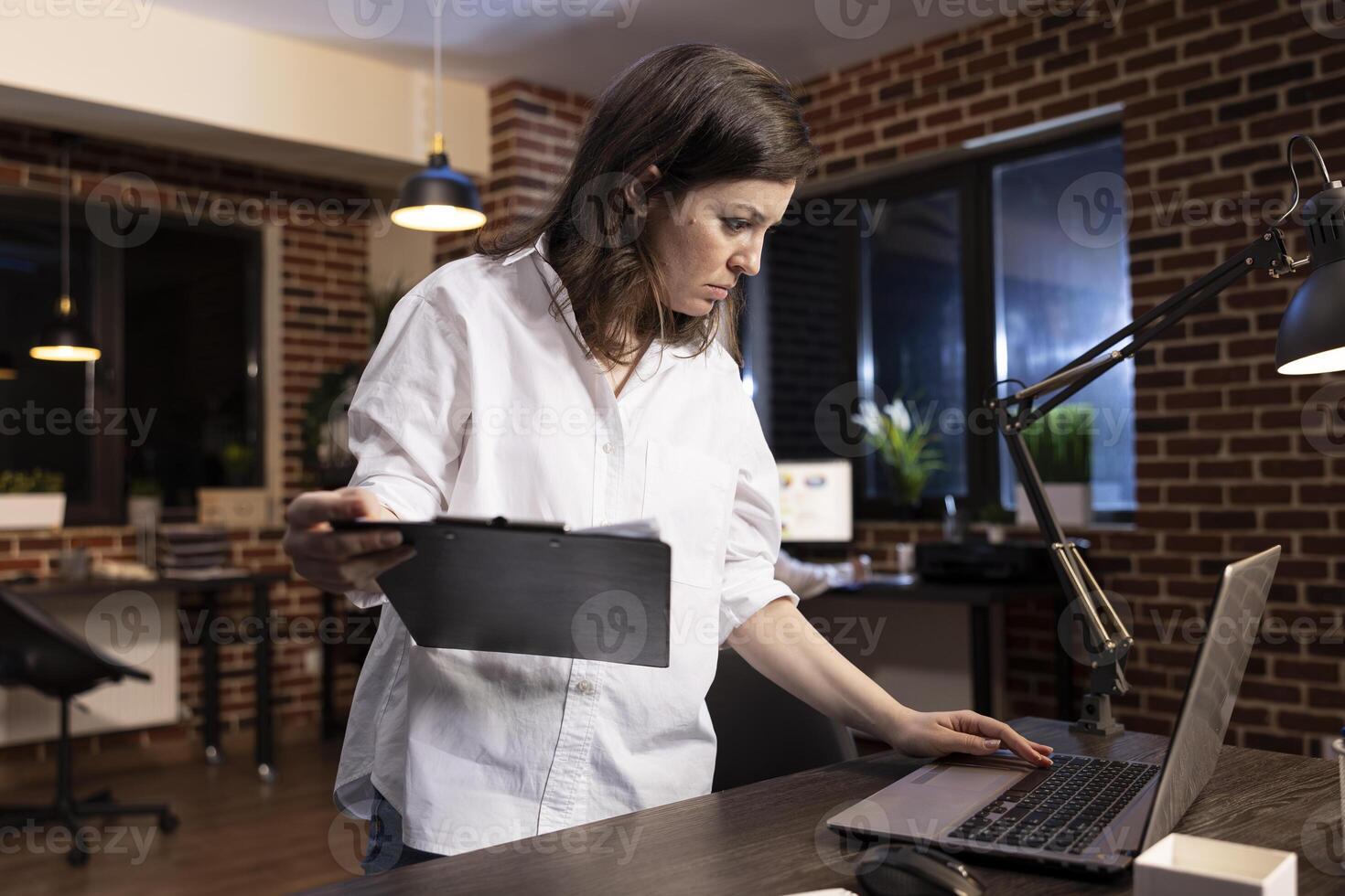 Late at night in a brick wall office, a focused businesswoman stands at her desk, comparing documents on clipboard and laptop. She carefully examines data, ensuring accuracy before making decisions. photo