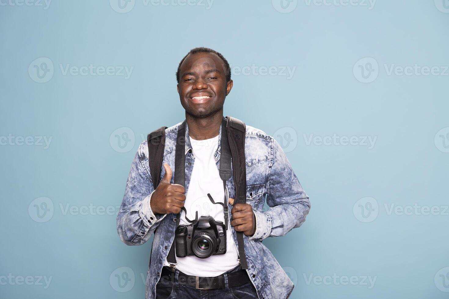 Cheerful black photographer enthusiastically gestures a thumbs up, ready for his next project. African american cameraman smiles brightly, equipped with a backpack and DSLR camera. photo