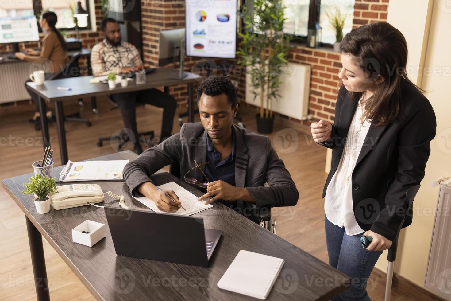 Businesswoman with a crutch collaborating with a colleague in wheelchair, sharing insights on marketing strategies. Diverse entrepreneurs with disabilities having a discussion in brick wall workspace. photo