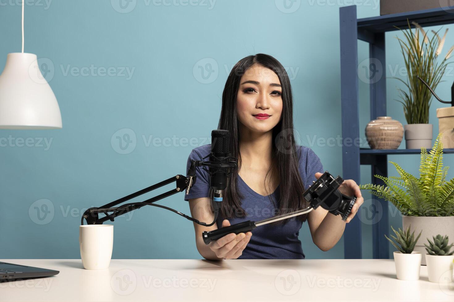 In a professional studio setup, female blogger grasping a camera tripod and talking into a microphone during her livestream. Young asian woman, seated at her desk, reviews content creation equipment. photo