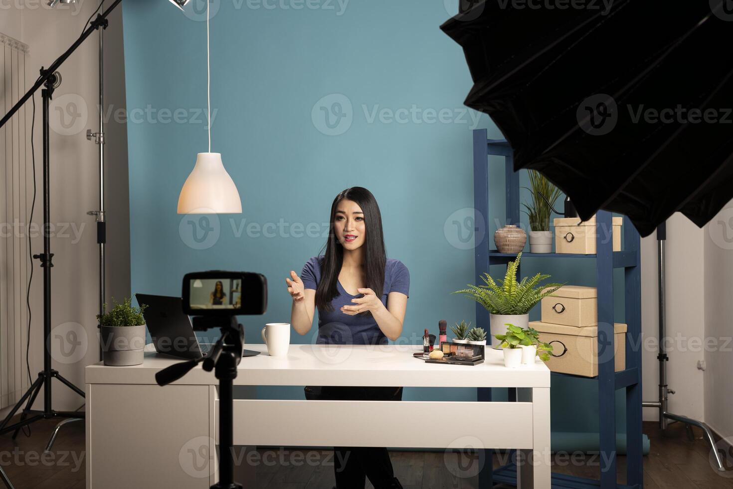 Young asian blogger recording a cosmetic review in a studio with a camera on a tripod. She sits at a desk with makeup products, using professional lighting and a blue background. photo