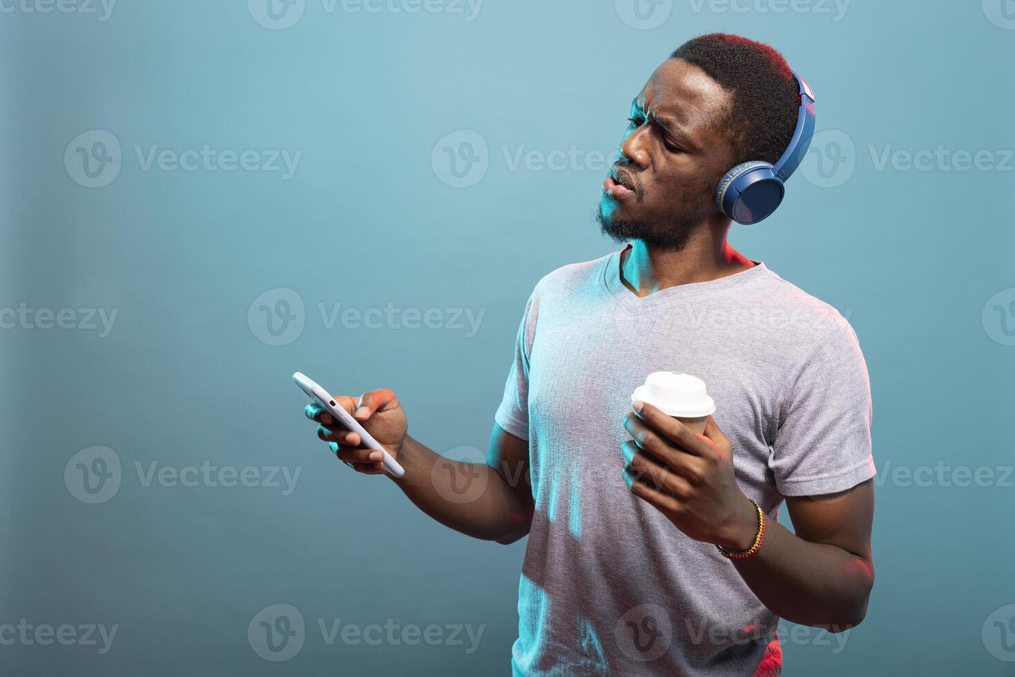Man wearing blue headphones, holding his coffee and checking his phone with a shocked expression. African american guy with disposable cup reacting in surprise to startling news on his mobile device. photo