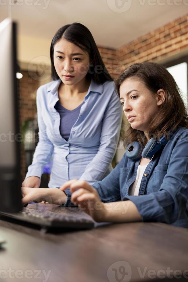 Two diverse women in modern office engaged in a creative discussion, one typing on the keyboard as the other reviews the work on the screen. Asian and caucasian colleagues collaborating on a project. photo