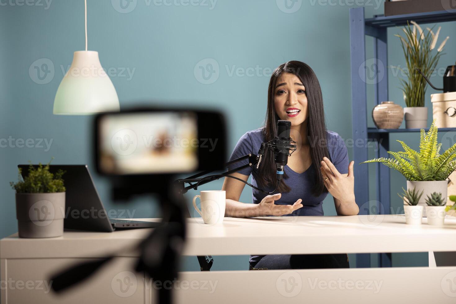 Young female vlogger seated at desk, talking into her podcast mic about marketing strategy for her channel. Asian woman using a camera on tripod, creating lifestyle content for her online audience. photo