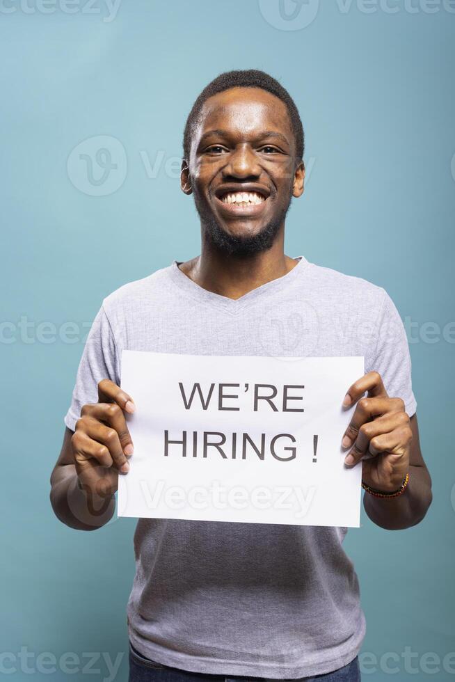 African American businessman holding a job hire sign and standing against a blue background with excitement. Cheerful male individual presenting open recruitment opportunities for candidates. photo