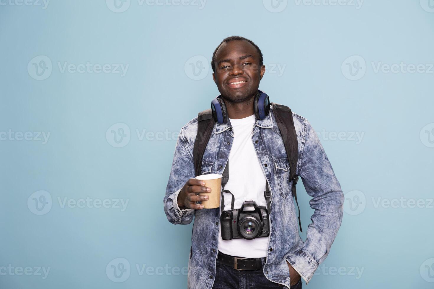 Portrait of male photographer with DSLR and headphones around his neck, confidently posing with one hand in pocket. Black man in casual clothing standing with a cup of coffee and looking at camera. photo