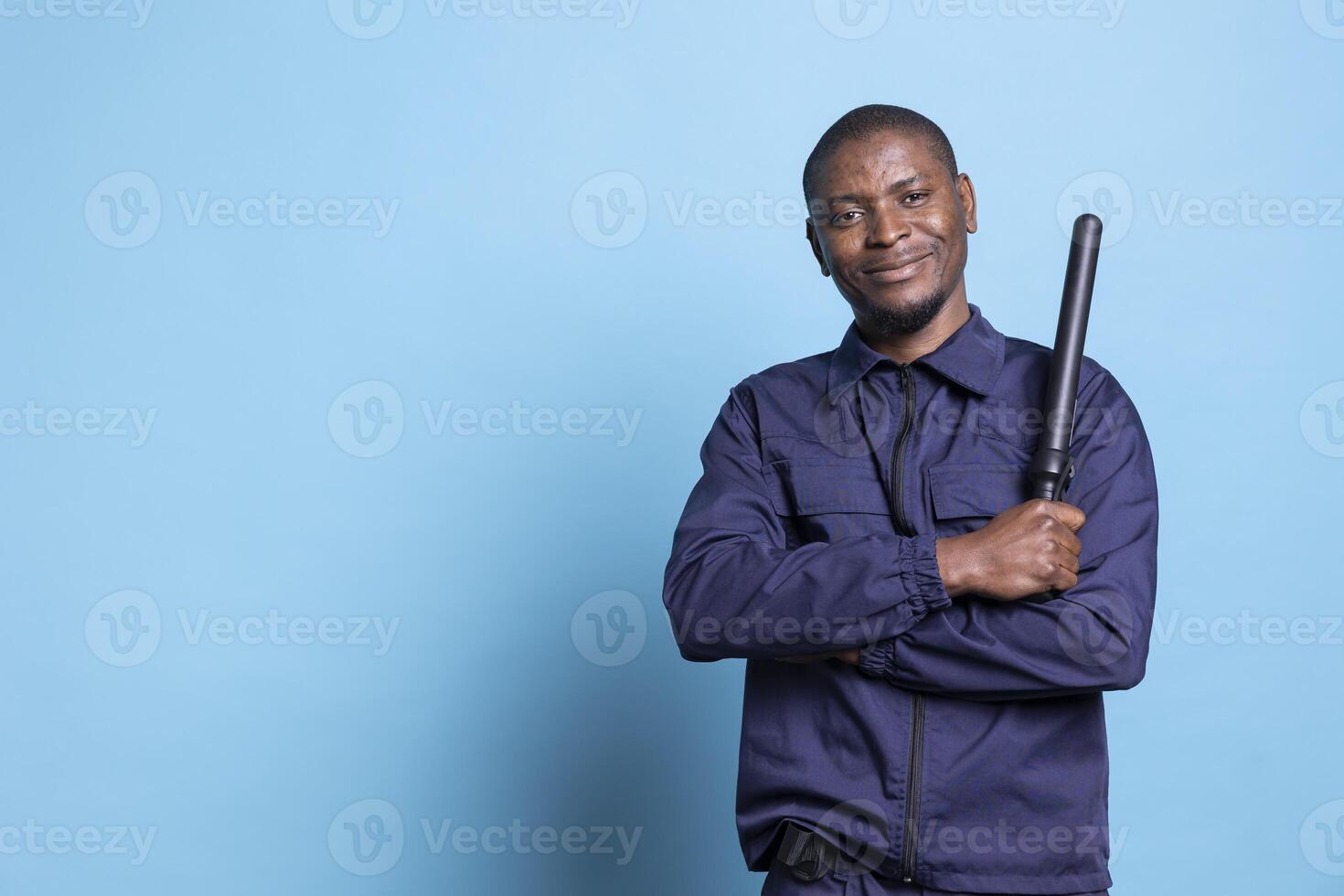 African american bodyguard posing with a baton and uniform in studio, showing confidence for his safeguarding duty. Skilled patrol officer standing against blue background on the job. photo