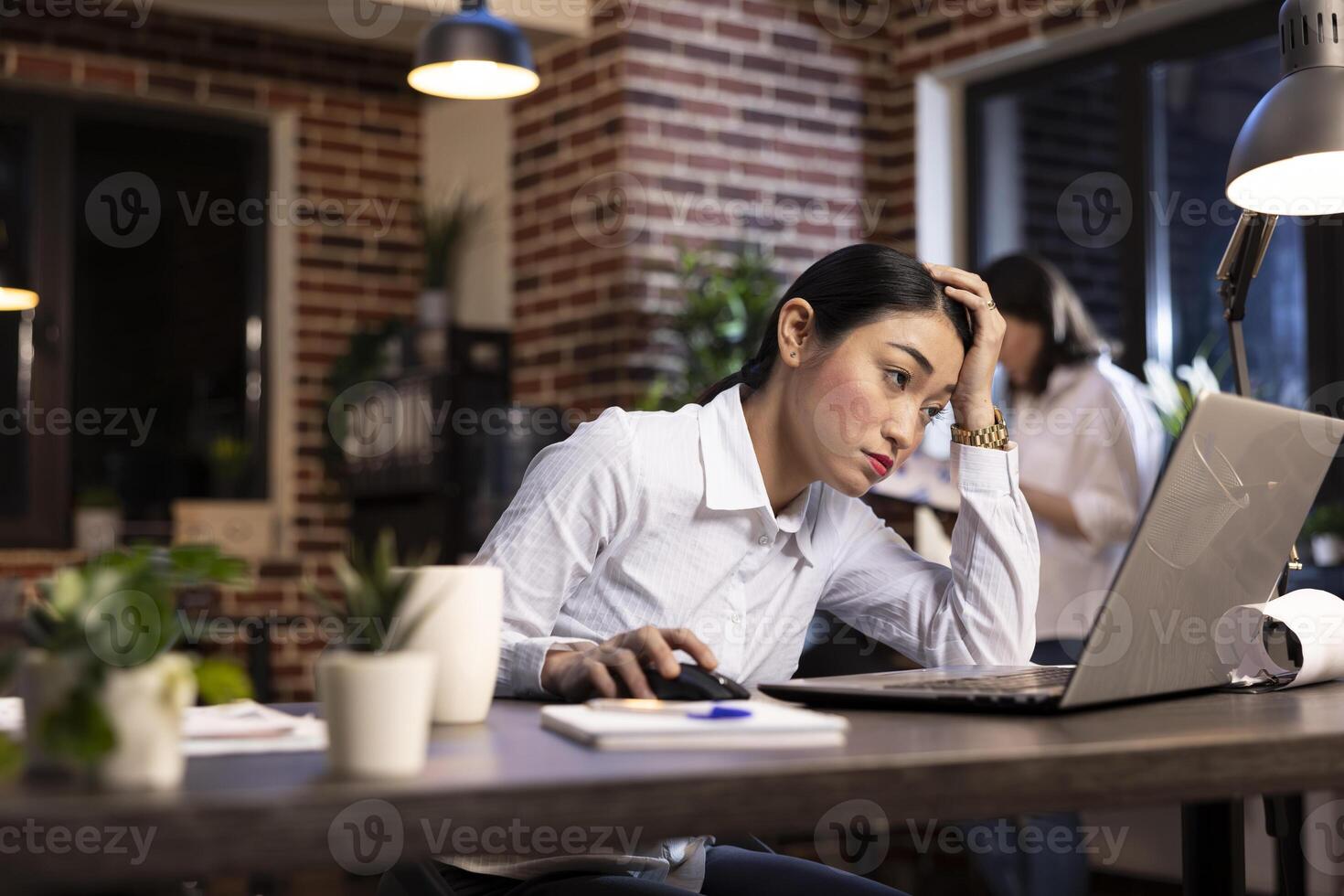 Tired asian businesswoman working late at her desk in a brick walled office, struggling to stay awake while scrolling through her laptop. Her coworker reviews documents in the background. photo