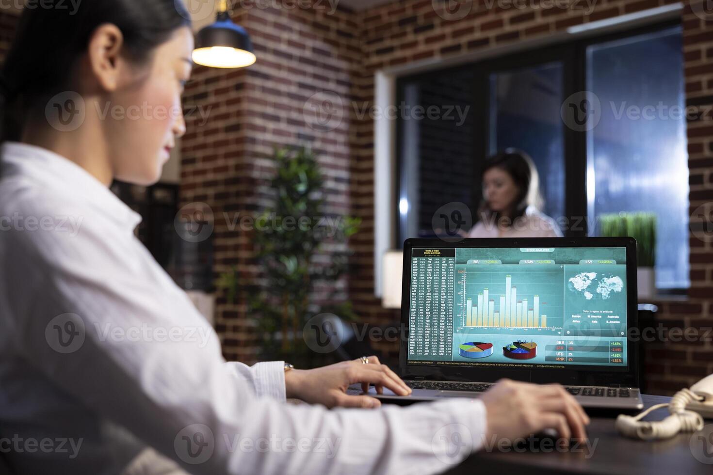 Female business analyst using her personal computer to examine marketing trends and track company performance in brick wall office. Asian employee holding her mouse as she scrolls through her laptop. photo