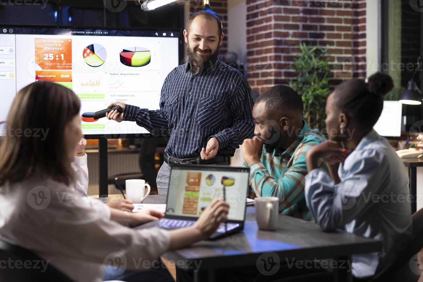 Caucasian man gives business presentation as coworkers analyze financial data on screen during night meeting in modern workspace. Laptop, documents and teamwork drive the strategic planning session. photo