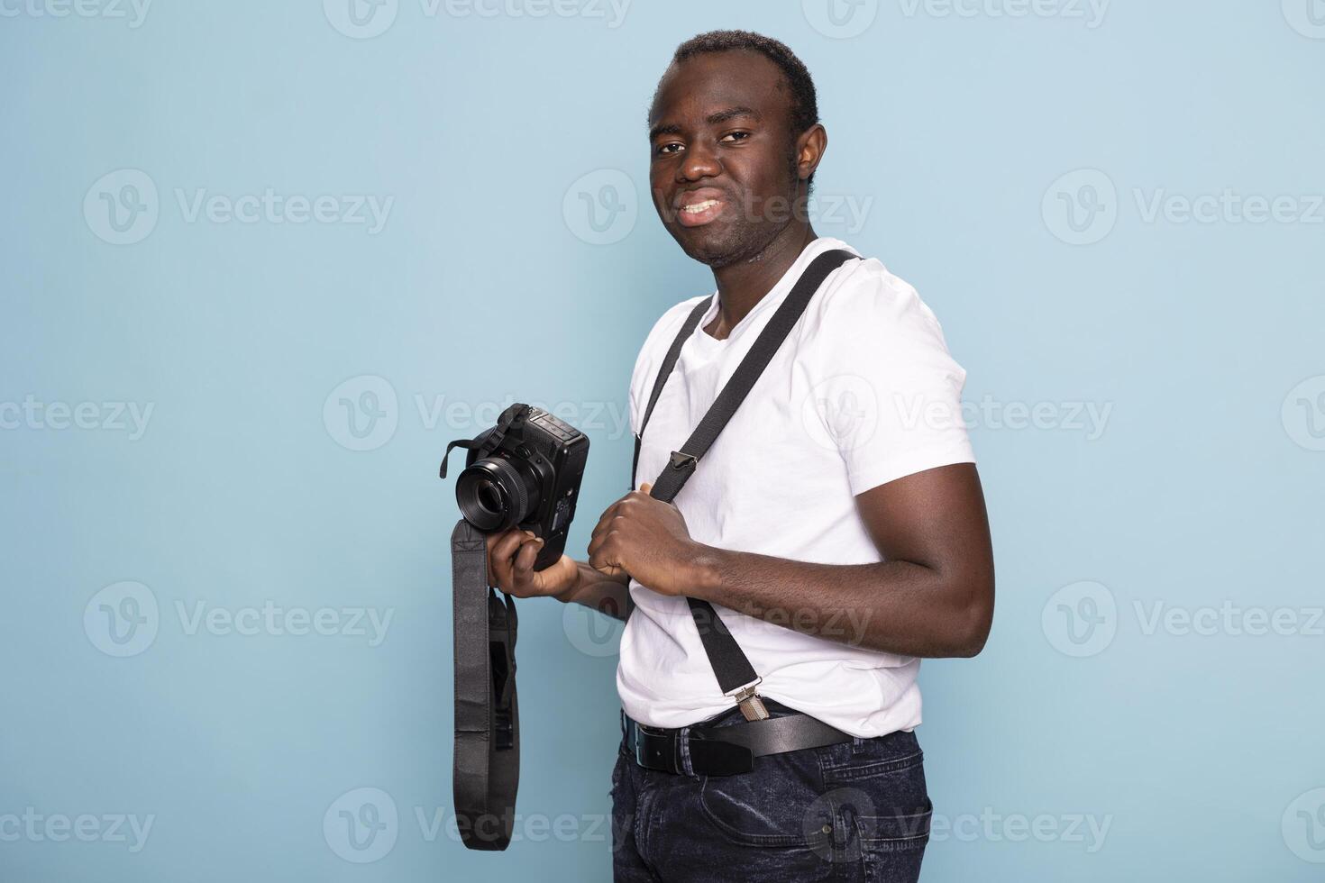 Black man dressed in a casual outfit with suspenders poses with his camera in front of a blue backdrop. Portrait of a male photographer, ideal for advertising, branding, and promotional use. photo