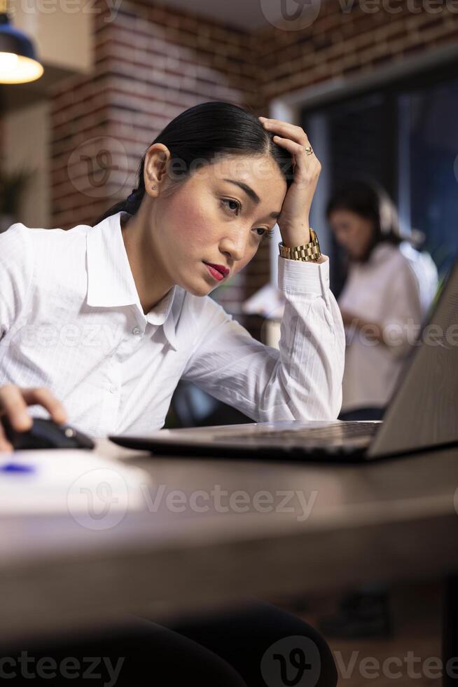 Asian female manager showing signs of fatigue while working late at her desk. Stressed businesswoman leans on one hand and looks at her laptop, as her blurred coworker reviews documents behind her. photo