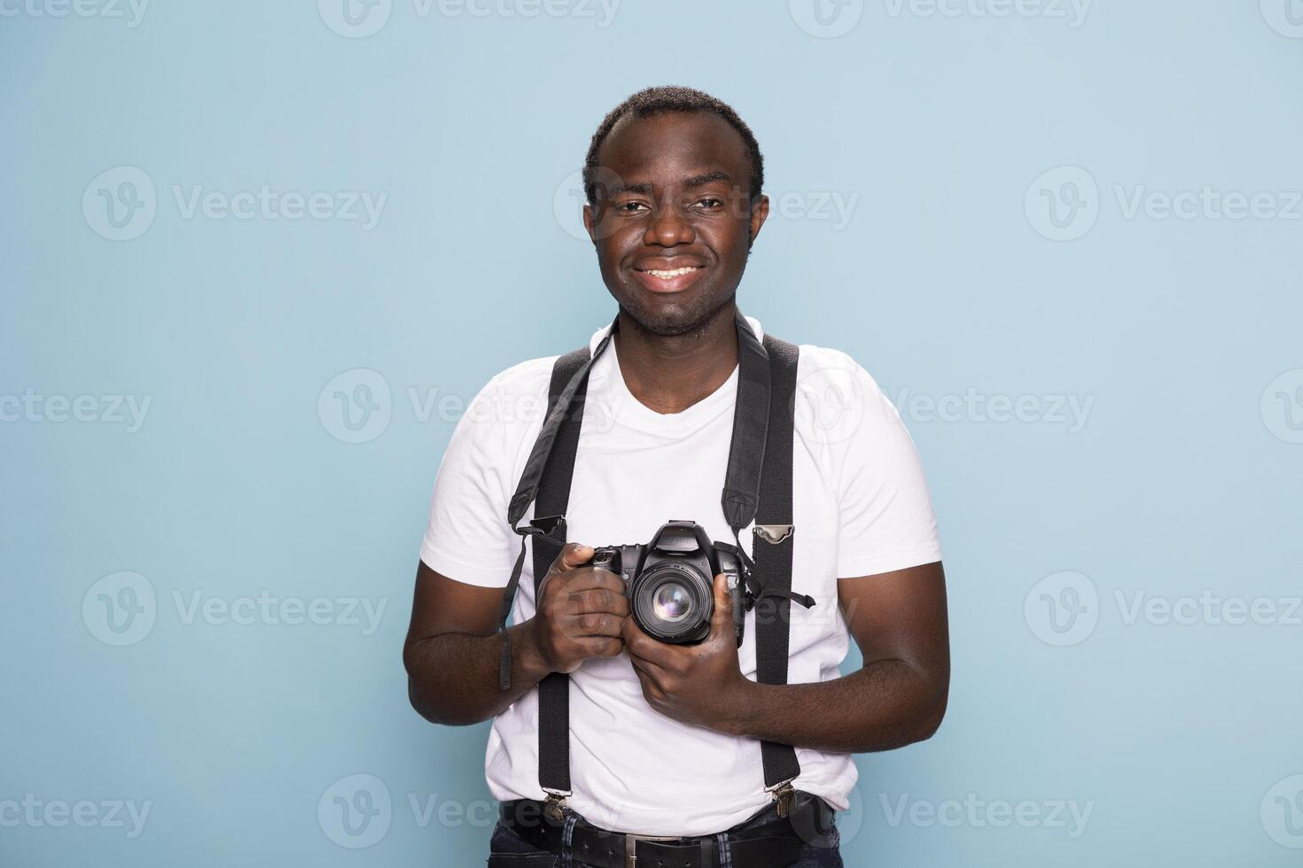 Cheerful male photographer portraying his creative side in a studio setting. Black man wearing stylish suspenders, he confidently holds his camera and poses against an isolated blue backdrop. photo