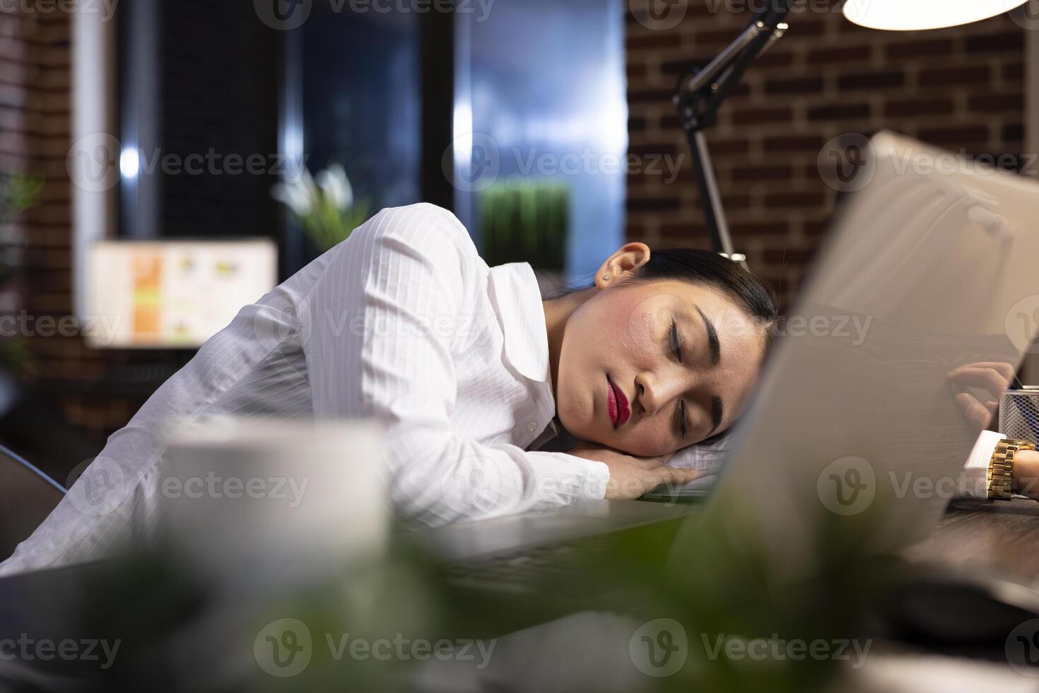 Overworked corporate manager sleeping at her office desk, taking a break during her night shift. Her laptop sits on the desk, highlighting the fatigue of long working hours in a modern workspace. photo