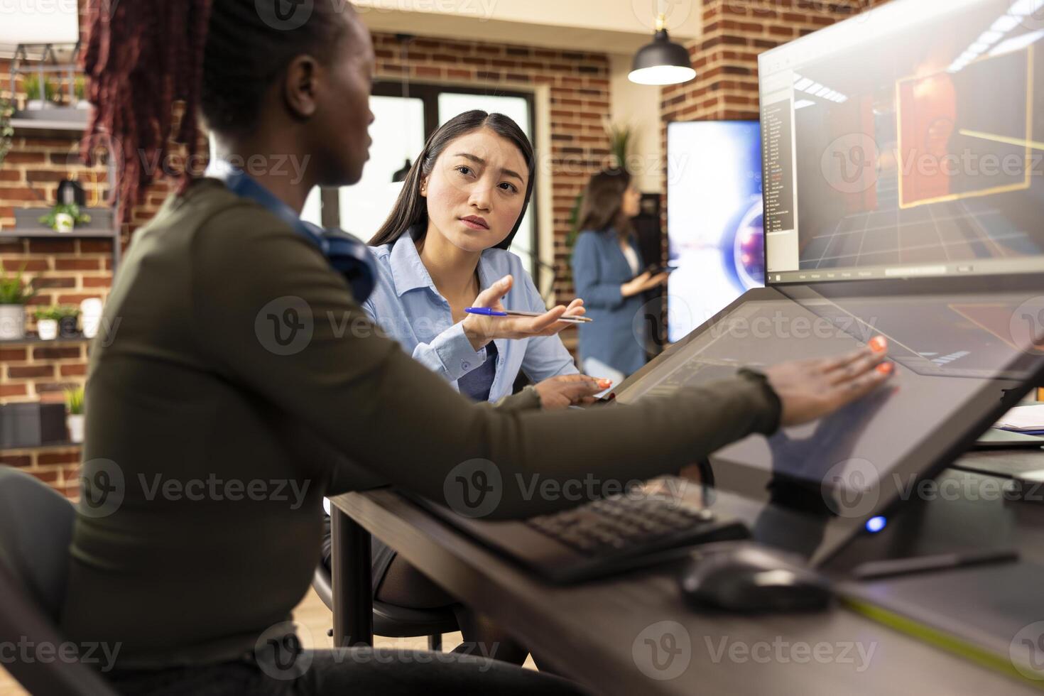 Female creative team leader with a pen in hand, questioning her designer working on the touchscreen monitor. Asian manager looking at her employee, asking for progress updates on product designs. photo